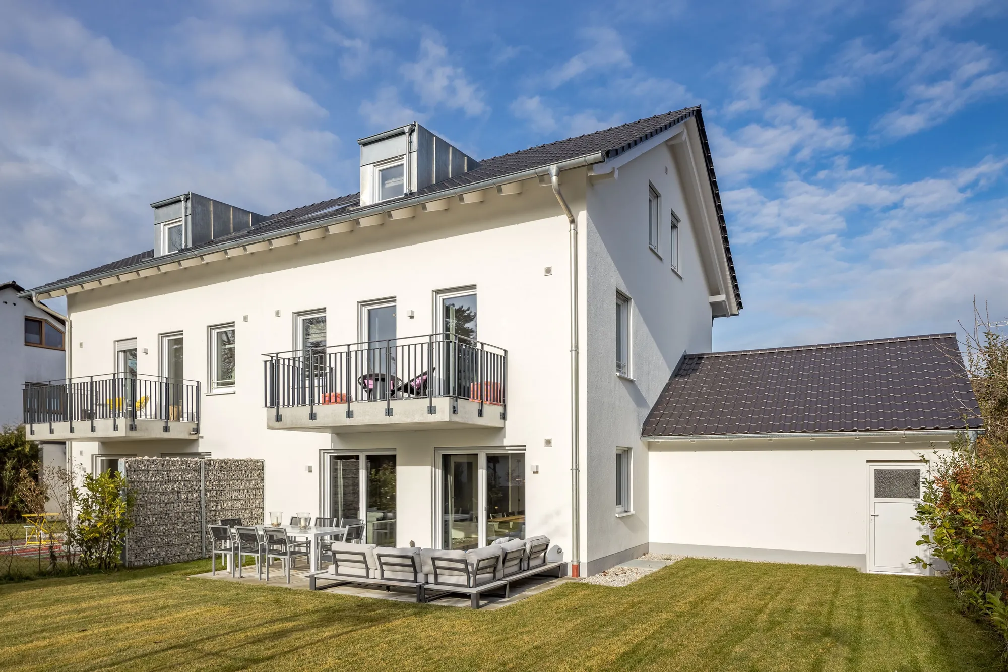 Helles Mehrfamilienhaus mit Balkonen, Gauben und Garten unter blauem Himmel